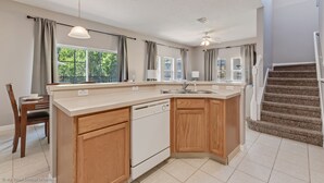 A modern kitchen with a large island, wooden cabinets, and a built-in dishwasher. There is a dining area with a table and chairs, and a staircase leading to another level.