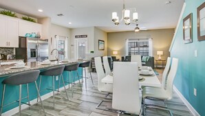 A modern kitchen with a central island, bar stools, and a dining area with white chairs and a table. - Slice Way Villa Champions Gate 1 (Davenport)