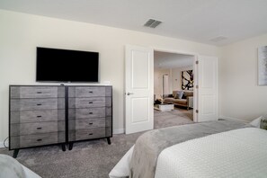 Una camera da letto con una grande TV a schermo piatto montata sopra una cassettiera grigia, un letto con coperta grigia e una porta aperta che dà su un'altra stanza con un divano e un tavolino.