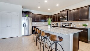 A modern kitchen with dark wood cabinets, stainless steel appliances, a granite countertop, and bar stools. - Bengal Court Villa Windsor at westsdie 8805 (Kissimmee)