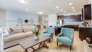 A spacious living area with a sofa, two armchairs, and a coffee table. A kitchen with dark cabinets and a countertop is visible in the background. - Bengal Court Villa Windsor at westsdie 8805 (Kissimmee)