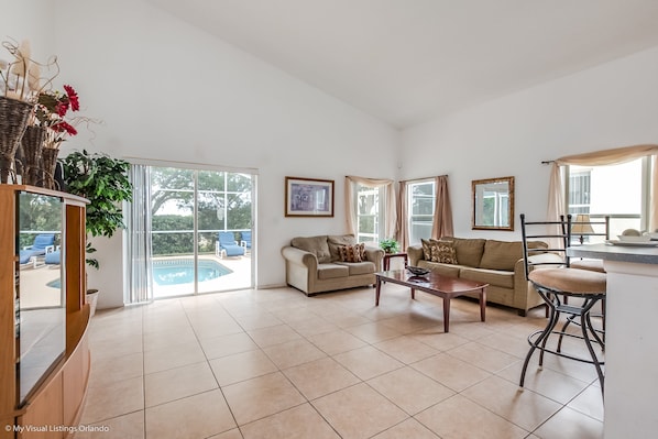 A spacious living room with a beige sofa, a coffee table, and a bar area. There are large windows with curtains, a pool visible outside, and a framed picture on the wall.