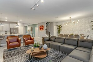 Una sala de estar moderna con un sofá seccional, sillones de cuero y una mesa de centro. Una escalera conduce a un comedor con una araña.