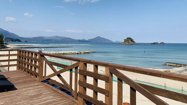A wooden deck leading to a beach with clear blue water, a distant island, and mountains in the background. - Beach Side Village Familiar - Hostel (Takahama)