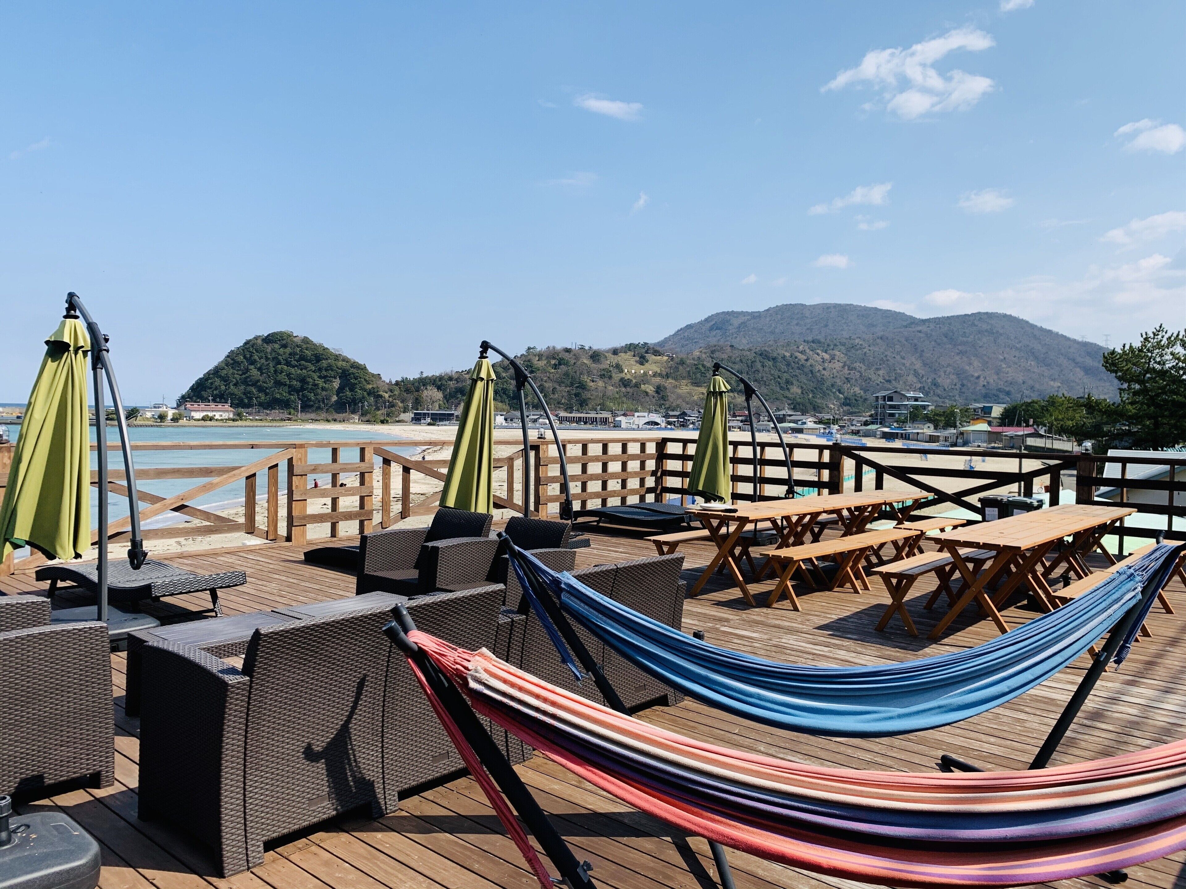A wooden deck with wicker furniture, a hammock, and green umbrellas overlooking a beach and mountains.