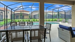 Ein überdachter Patio-Bereich mit einem Glastisch und Stühlen, ein Korbsessel mit Kissen und Blick auf einen Swimmingpool sowie Häuser im Hintergrund.