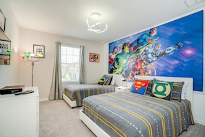 A bedroom with two beds, a large superhero-themed mural, and a desk with a lamp.