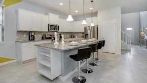 A modern kitchen with white cabinets, a granite countertop, stainless steel appliances, and a central island with bar stools. - Bengal Drive Villa Windsor At Westside 8917 (Kissimmee)
