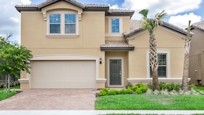 A two-story house with a beige exterior, a white garage door, and a small front yard with a palm tree and shrubs. - Bengal Drive Villa Windsor At Westside 8917 (Kissimmee)