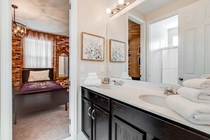 A modern bathroom with a large mirror, a double sink, and a bed with a brick wall backdrop.