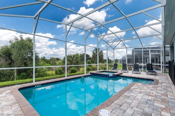 Un área de piscina con cerramiento de vidrio, solárium con baldosas y vista a la vegetación circundante.