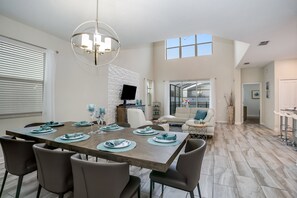 A spacious dining area with a wooden table set for six, surrounded by modern chairs. A large chandelier hangs from the ceiling, and a flat-screen TV is mounted on the wall. - Amazing Storey Lake 5 Bedroom 5 Bathroom 4778kcc (Kissimmee)