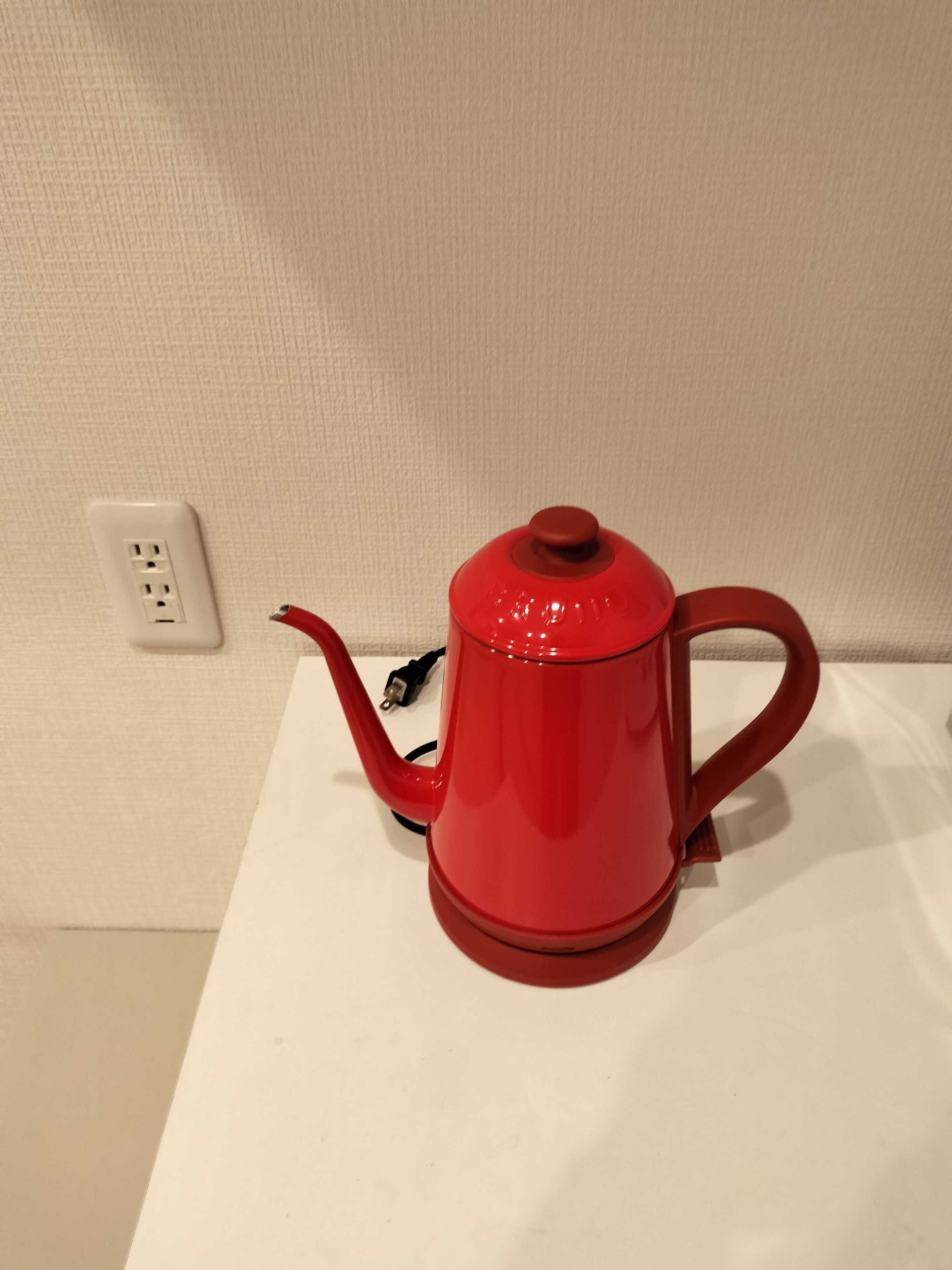 A red kettle on a white table against a textured wall with a power outlet.