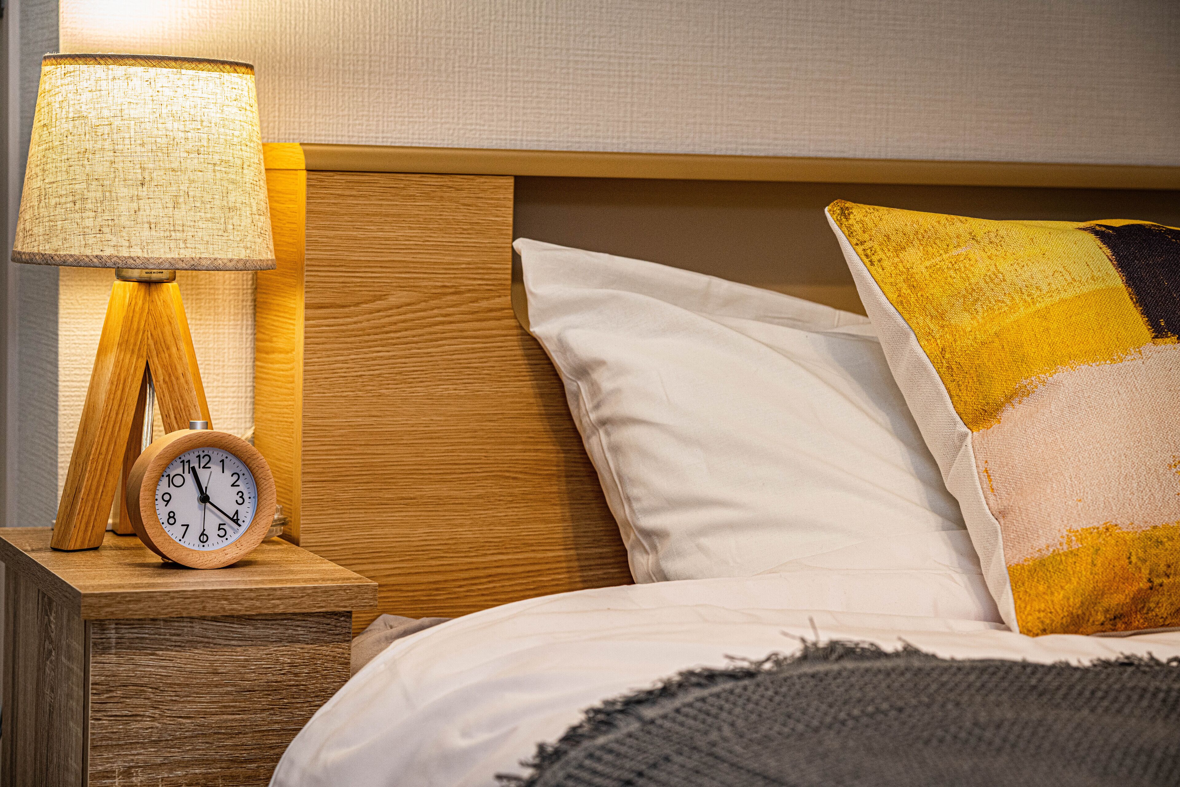 A wooden bedside table with a clock and a lamp, a bed with white and multicolored pillows, and a textured wall in the background.