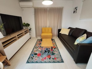 A living room with a flat-screen TV, a yellow sofa, a dark sofa, a floral rug, and a white cabinet.