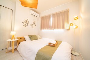 A neatly arranged hotel room with a bed, bedside table, lamp, and a wall-mounted light fixture. - Tachibana House (Tokyo)