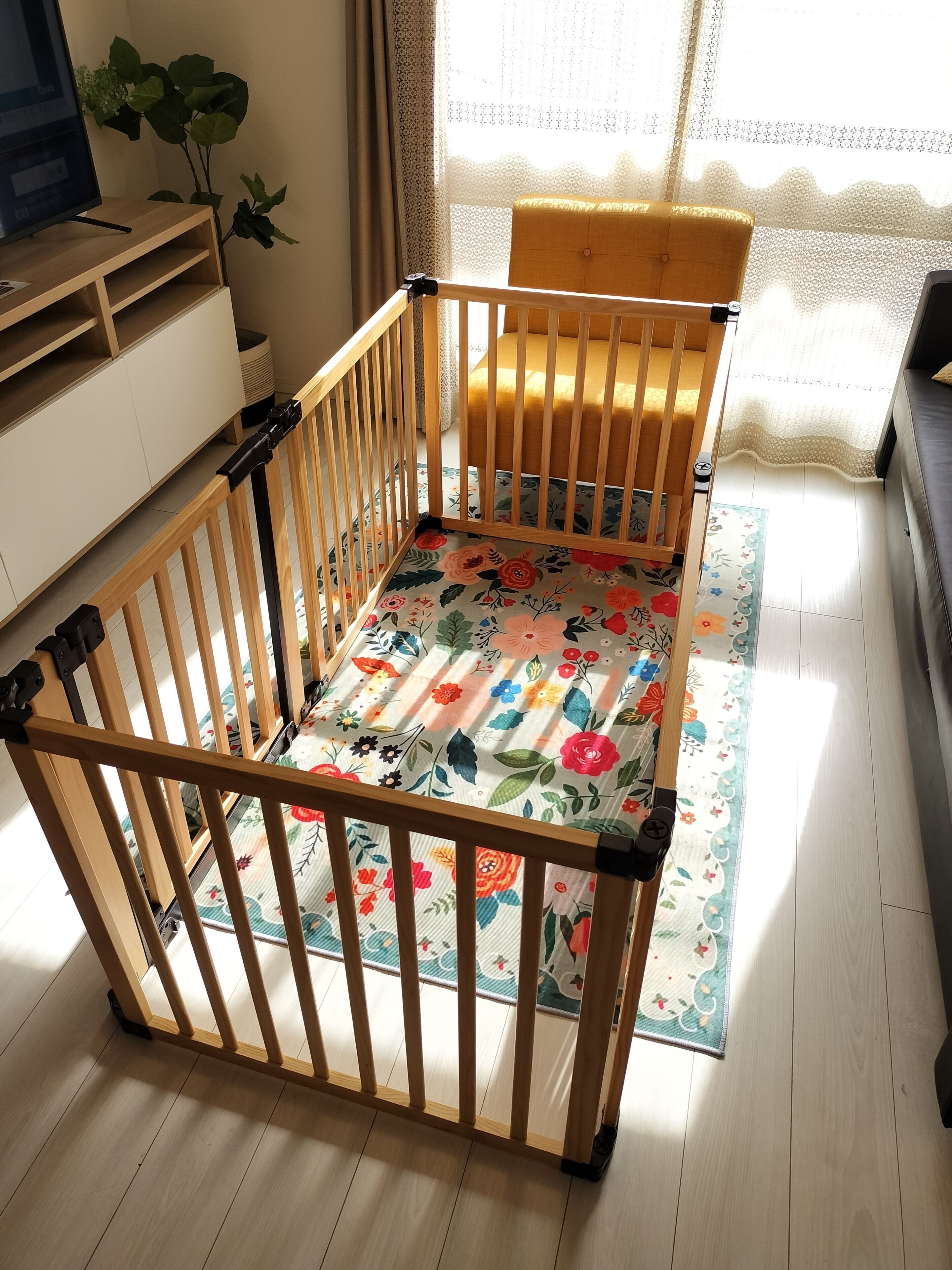 A wooden crib with a floral mattress, a wooden floor, a window with sheer curtains, and a plant.