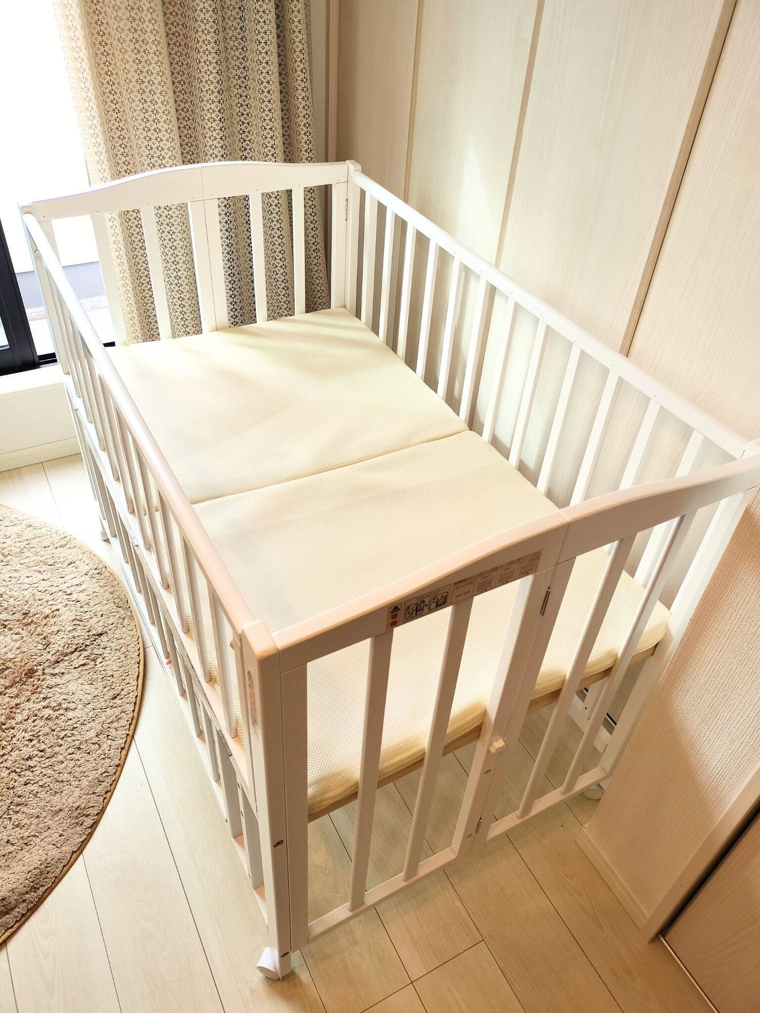 A white baby crib with a beige mattress, positioned in a room with wooden flooring and a window with curtains.