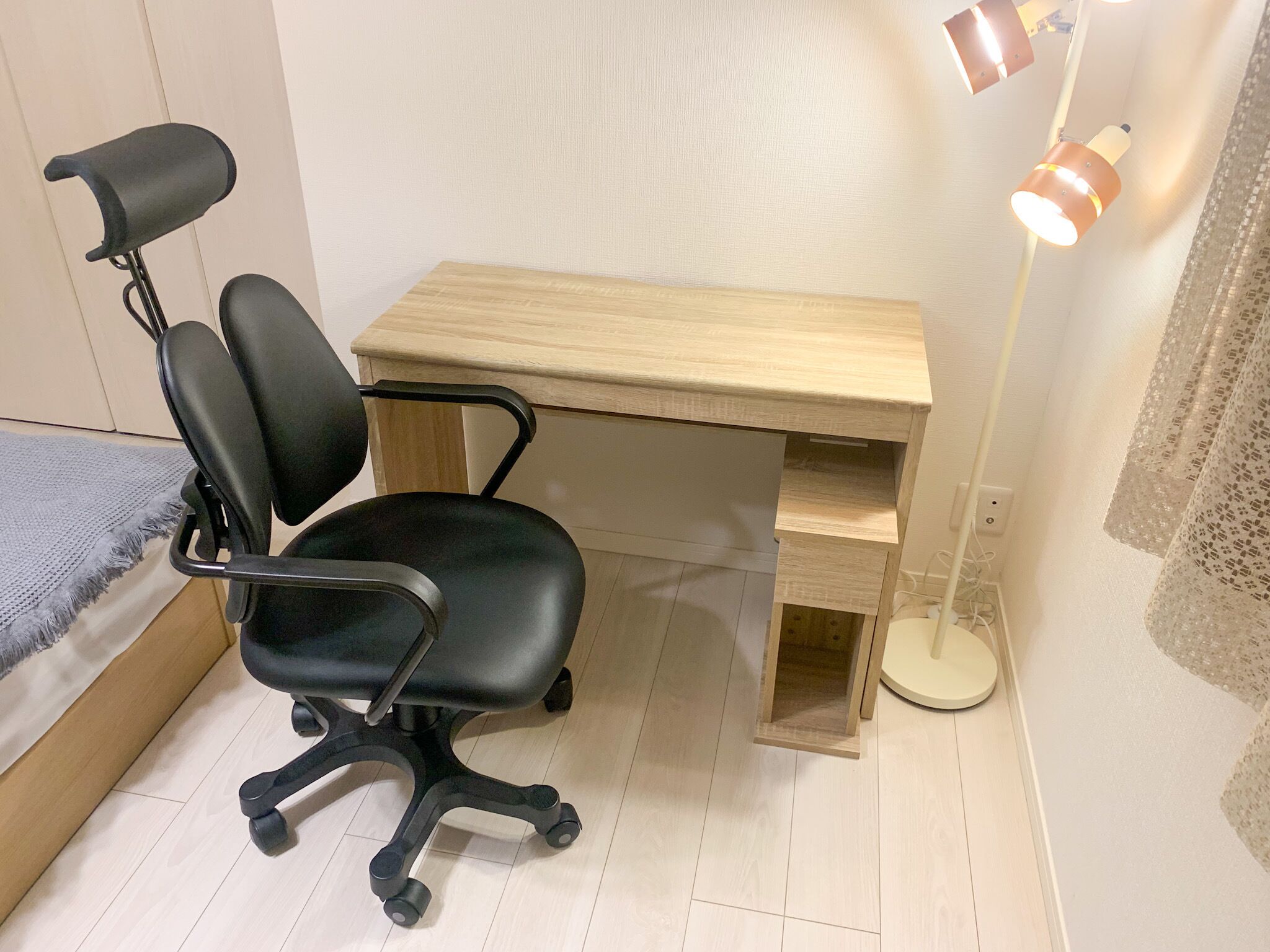 Una habitación pequeña y ordenada con un escritorio de madera, una silla de oficina negra, una mesita de noche y una lámpara de pie.