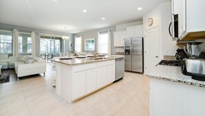 A modern kitchen with white cabinetry, a central island, stainless steel appliances, and a dining area with a white sofa. - Oakshaddows Villa Windsor At Westside (Davenport)