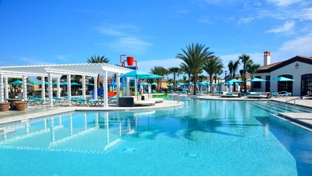 A swimming pool with lounge chairs and umbrellas, surrounded by palm trees and a covered area with a water slide. - Sydney Villa Windsor At Westside 1 (Kissimmee)