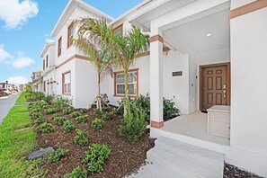 A two-story house with a white exterior and brown trim, a front porch, and a well-maintained garden with various plants and a palm tree.