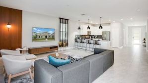 A spacious living area with a grey sectional sofa, a small round coffee table, and a white armchair. A kitchen with modern appliances and bar stools is visible in the background. - Target Villa Story lake (Kissimmee)