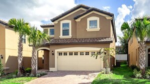 Een tweelaags huis met een beige gevel, een bruin dak en een witte garage. Voor het huis staan palmbomen en een verzorgd gazon.