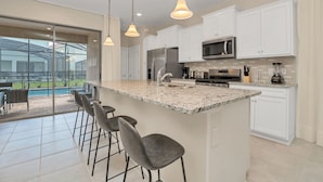 A modern kitchen with white cabinets, a large island with bar stools, stainless steel appliances, and a view of a swimming pool. - Misty Villa Champions Gate 5469 (Davenport)