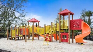 A playground with a yellow and red play structure, including slides and climbing areas, set in a sandy area with trees in the background. - Oakmoss Drive Villa Solterra 1 (Davenport)