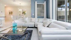A modern living room with a white sectional sofa, a glass coffee table, and a blue rug. There is a dining area in the background with a white table and chairs. - Romeo Circle Villa Story Lake 2 (Kissimmee)