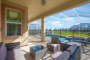 Uma área externa coberta com mobiliário de vime, uma mesa com tampo de vidro e vista para uma piscina e algumas casas.