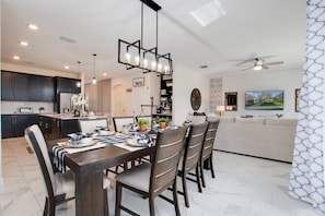 Uma cozinha moderna com armários escuros, um espaço de jantar com mesa e cadeiras de madeira, e uma sala de estar com sofá e televisão.