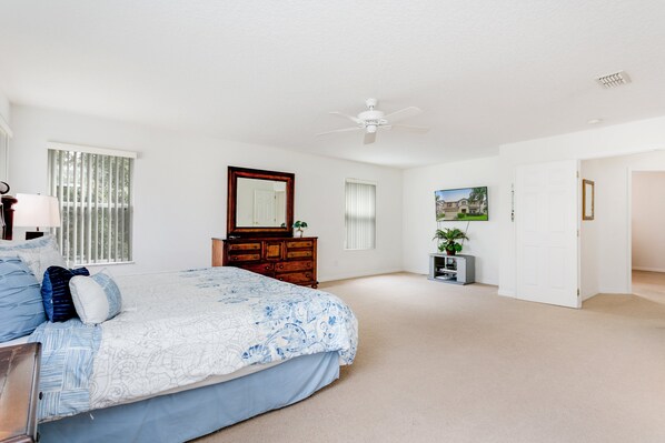 A spacious bedroom with a large bed, a dresser, a ceiling fan, and a door leading to another room.