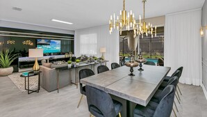 A modern dining area with a large table, chairs, and a chandelier. There is a kitchen area with a dining table and chairs in the background.