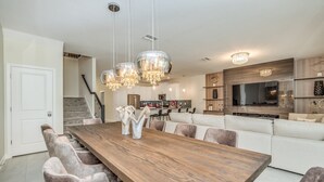 Un vaste espace de salle à manger avec une table en bois et des chaises rembourrées, une cuisine équipée d’appareils électroménagers modernes et un grand téléviseur fixé au mur.
