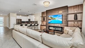 Un salon moderne doté d’un grand téléviseur à écran plat, d’un canapé d’angle beige, d’un coin repas avec des tabourets de bar et d’une cuisine en arrière-plan.