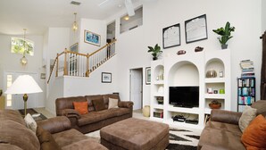 A spacious living room with a staircase, leather sofas, a flat-screen TV, and a bookshelf.