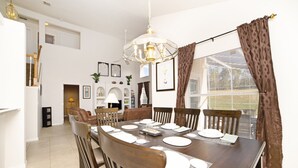 A dining area with a large wooden table set for six, surrounded by wooden chairs. There is a chandelier above the table, and the room has white walls and a large window with curtains.