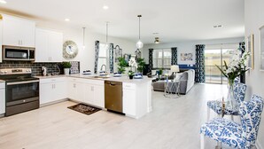 A modern kitchen with white cabinets, a built-in oven, and a dishwasher. There is a dining area with blue patterned chairs and a table.