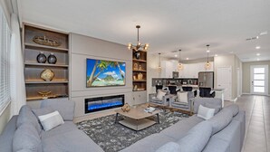 Un salon moderne avec un canapé d’angle gris, un téléviseur à écran plat, une cheminée et une cuisine en arrière-plan.