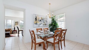 A dining area with a glass table, wooden chairs, and a chandelier. There is a large abstract painting on the wall and a potted plant near the window. - Pine Villa Winwood Bay (Davenport)