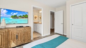 A bedroom with a bed, a TV on a wooden cabinet, a bathroom with a sink and mirror, and a door leading to another room.