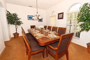 A dining room with a wooden table set for six, surrounded by six chairs. There are two potted plants, a chandelier, and framed pictures on the walls.