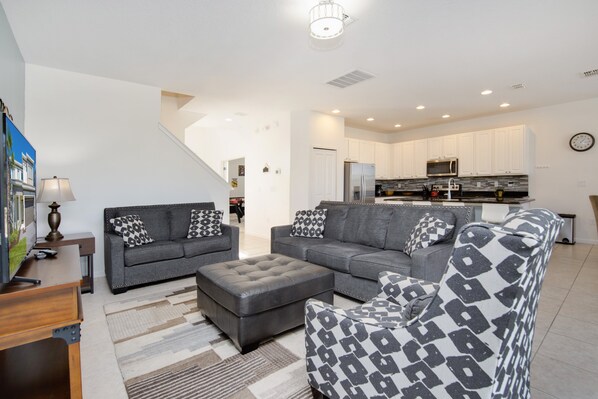 A modern living room with a grey sofa set, a matching ottoman, and patterned throw pillows. A wooden coffee table is in front of the sofa. The room has a staircase leading to an upper level. - Callistoga Villa Sonoma 3 (Kissimmee)