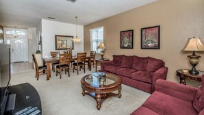 A living room with a red sofa, a coffee table, and a TV. There is a dining area with a table and chairs. - Oxford Drive Villa Highlands Reserve (Davenport)
