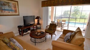 A living room with a sofa, armchair, and coffee table, a TV, and a view of a swimming pool. - 7783tb Windsor Hills Resort 5 Bed 5 Bath (Kissimmee)