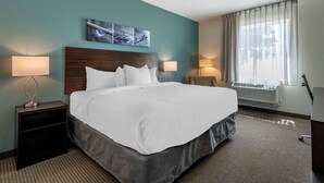 A hotel room with a large bed, two bedside tables with lamps, a wall-mounted artwork, a window with sheer curtains, and an air conditioning unit.