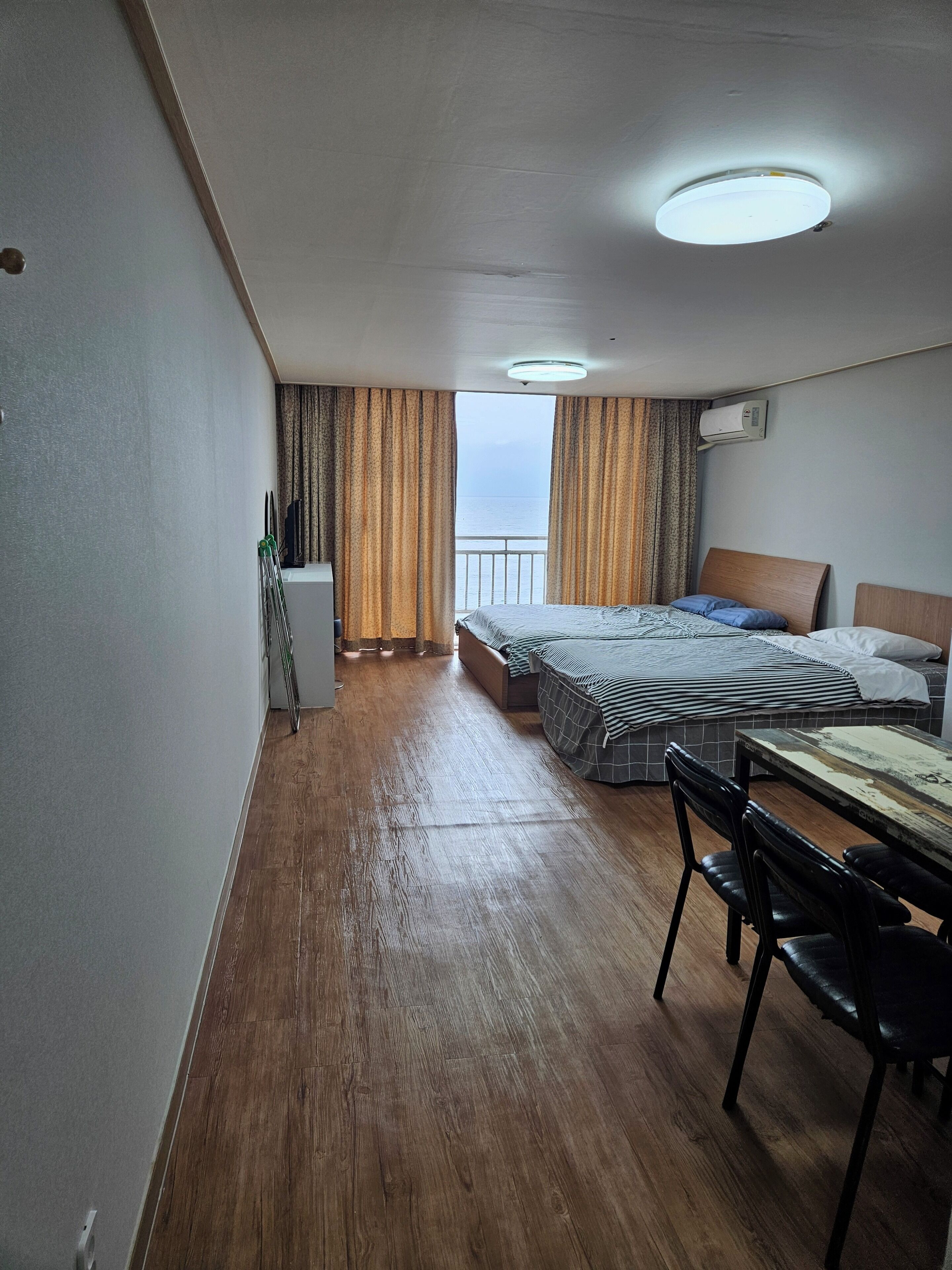 A hotel room with a wooden floor, a bed with a striped bedspread, a desk with a chair, and a view of the sea through curtains.