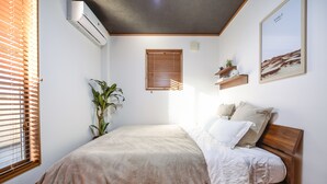 Una camera da letto con un letto, una pianta in vaso, un condizionatore d'aria a parete, una finestra con persiane e un quadro appeso al muro.
